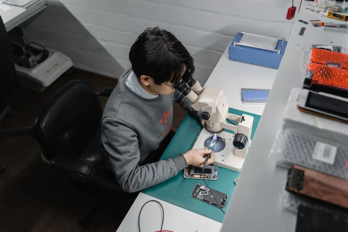 Un tecnico esamina uno smartphone aperto al microscopio sul banco di lavoro.